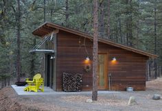 a small cabin in the woods with two yellow chairs and a firewood log holder