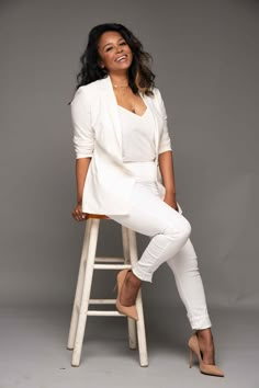 a woman sitting on top of a wooden stool wearing white pants and blazer jacket