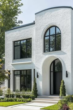 a white brick house with arched windows and black shutters on the front door is surrounded by greenery