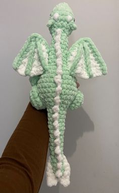 a crocheted dragon stuffed animal being held by a person's hand in front of a gray wall