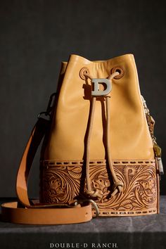 FOR THE LOVE OF LEATHER Double D Ranch is getting in the handbag game! This luxury leather bucket bag is hand-tooled with classic western scrolling and finished with darling DDR dangles and a slide cinch. Cool, classic, and collectible – these are a limited-edition run! artist: Double D Ranch materials: Argentinean Leather embellishments: hand-tooled, custom Double D findings, contrast lacing measurements: 12" x 8 1/4" x 9.5" strap: adjustable 41" -45" weight: 2.1 lbs contemporary style number: Leather Embellishments, Collar Tips, Double D Ranch, Earring Pins, Belt Jewelry, Leather Bucket Bag, Leather Bucket, Accessories Jewelry Necklace, Makeup Case