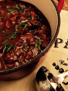 a pot full of stew sitting on top of a wooden table next to a spoon