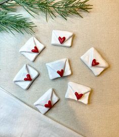 small white envelopes with red hearts are sitting on a table next to a pine branch