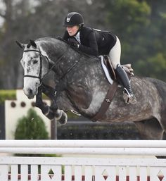 a woman riding on the back of a gray horse over an obstacle in a race