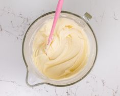 a mixing bowl filled with cream on top of a white marble countertop next to a pink plastic spoon