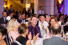 a group of people sitting at tables with wine glasses in front of them and clapping