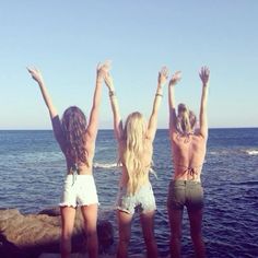 three young women standing on the edge of a cliff by the ocean with their arms in the air