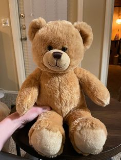 a brown teddy bear sitting on top of a wooden table next to a persons hand