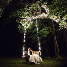 a bride and groom sitting on a bench under the trees at night with fairy lights