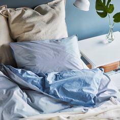 an unmade bed with blue sheets and pillows on it, next to a plant