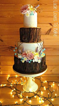 a three tiered wedding cake with flowers on the side and lights around it in front of a wooden wall