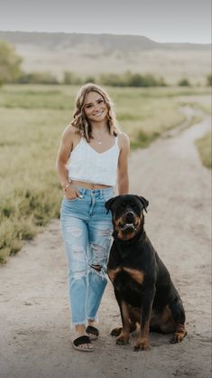 a woman standing next to a black and brown dog on a dirt road with grass in the background