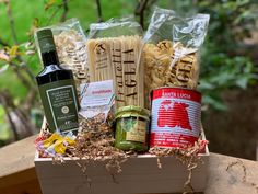 an assortment of pasta and wine in a wooden box on a table outside with trees