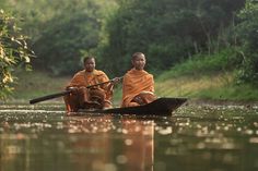 two men in orange robes are rowing a boat on the water with trees behind them