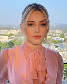 a woman with blonde hair wearing a pink shirt and tie looking at the camera while standing in front of a cityscape