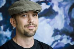 a man wearing a hat in front of a blue and white wall with an artistic painting behind him