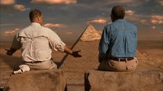 two men sitting on rocks looking at the pyramids