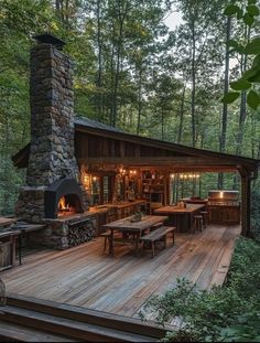 a wooden deck with tables and chairs next to a fire place in the woods at dusk