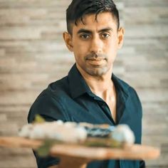 a man holding a wooden tray with food on it and looking at the camera while standing in front of a brick wall