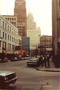 an old photo of people standing in the middle of a street with cars parked on both sides