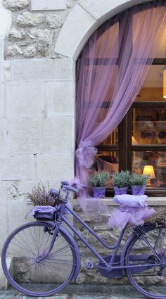 a purple bicycle is parked in front of a window with sheer curtains and lavender flowers