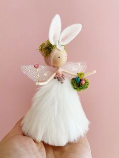 a hand holding a tiny white fairy doll with flowers on it's wings, against a pink background