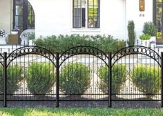 a black iron fence in front of a white house with green grass and bushes behind it