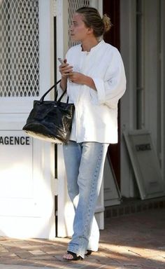 a woman in white jacket and jeans holding a black handbag while standing next to a building