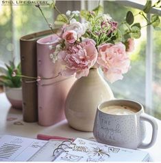 pink flowers in a vase next to a coffee mug and books on a table near a window