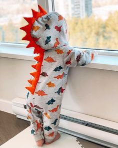 a toddler wearing a dinosaur onesie standing on a window sill looking out the window