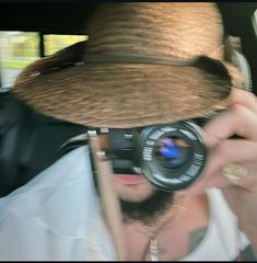 a man wearing a hat and holding a camera up to his face while sitting in a car