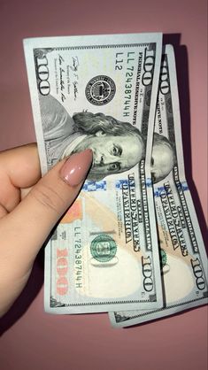 a woman's hand holding up two stacks of one hundred dollar bills in front of pink background