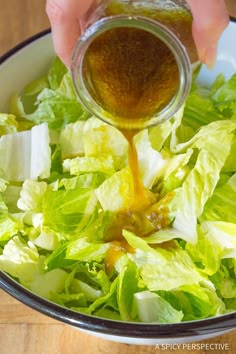 a person pouring dressing into a salad in a white bowl on a wooden table with lettuce and cheese