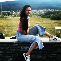 a woman sitting on top of a brick wall next to a lush green field with mountains in the background