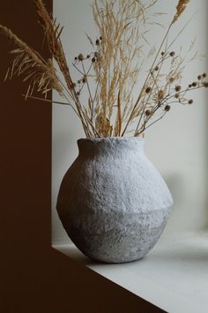 a white vase with dried plants in it on a window sill next to a window