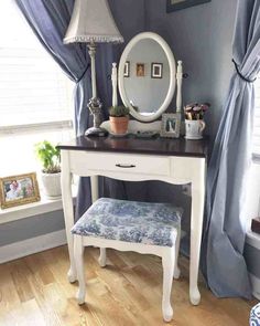 a white desk with a mirror, stool and lamp in front of blue drapes