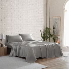 a bed with grey sheets and pillows in a white brick room next to a potted plant