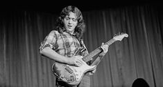 black and white photo of man playing guitar on stage with curtain in the back ground