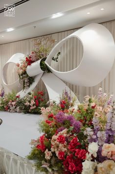 an arrangement of flowers on a table with a large white bow at the top and bottom