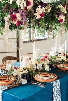 a blue table cloth topped with plates and place settings next to tall floral centerpieces