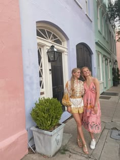 two women standing next to each other in front of a pink and blue building with potted plants