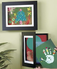 a person holding up some pictures in front of a wall with plants and paintings on it