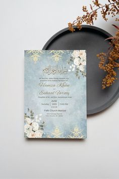 a blue and white floral wedding card on a plate next to some dried plant life