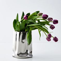 purple tulips are in a silver vase on a white surface with green leaves