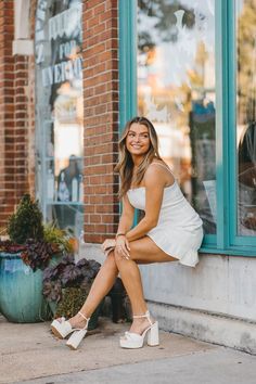 a woman sitting on the side of a building wearing high heels and a white dress