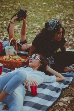 two women sitting on a blanket in the grass with picnic foods and drinks, while one woman holds up her cell phone