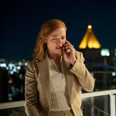 a woman talking on a cell phone while standing next to a balcony at night with city lights in the background