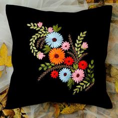 a black pillow with colorful flowers and leaves on the ground in front of some fall leaves