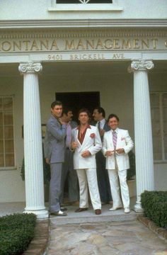 four men in white suits standing outside the front door of a building with columns and pillars