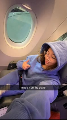 a woman sitting on an airplane seat wearing a blue hoodie and pointing at the window